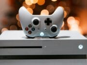A white Xbox One console with a matching controller, placed on a gray surface. The console has an illuminated power button, and blurred lights are in the background.