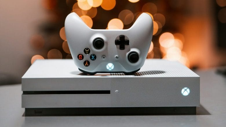 A white Xbox One console with a matching controller, placed on a gray surface. The console has an illuminated power button, and blurred lights are in the background.