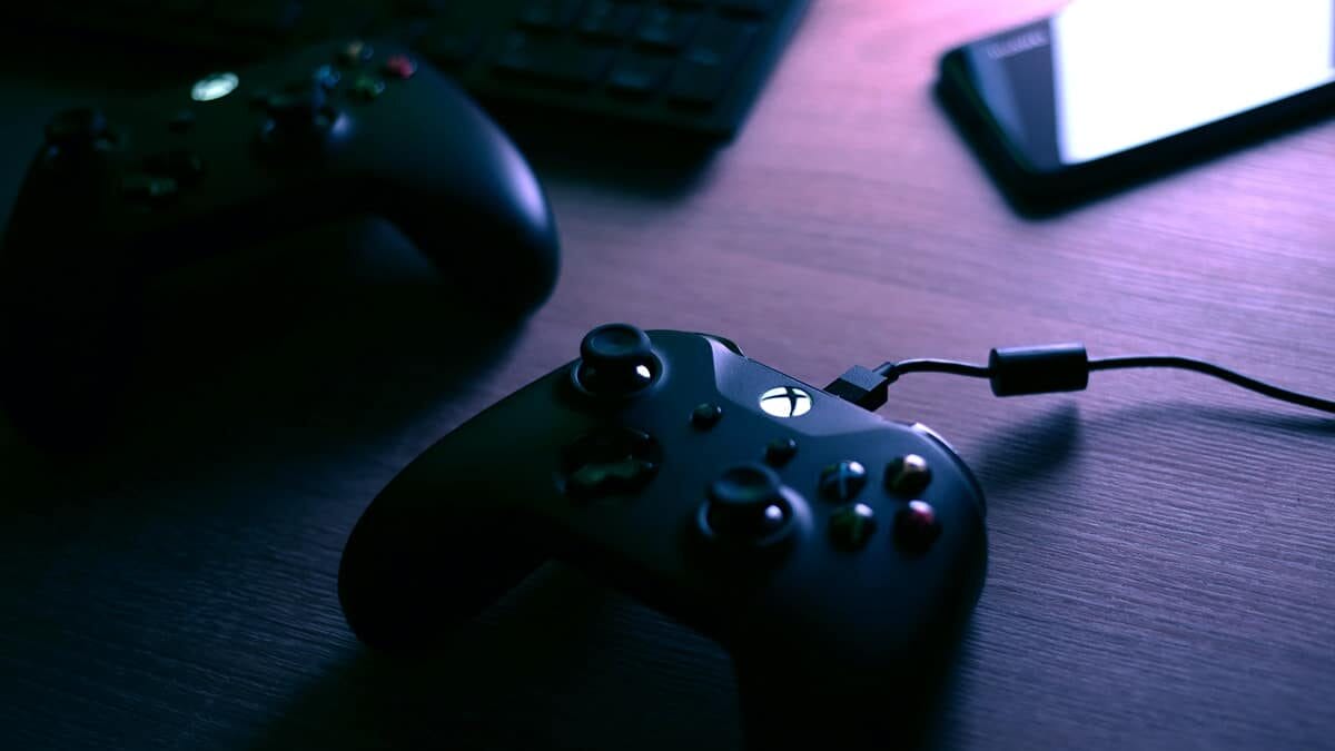 A black gaming controller is connected with a USB cable on a wooden surface, with another controller, a keyboard, and a smartphone nearby.