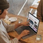 A woman in a beanie uses a laptop at a wooden table, pointing at the screen to show something to a person sitting across from her. they have plates and a glass of water on the table.