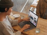 A woman in a beanie uses a laptop at a wooden table, pointing at the screen to show something to a person sitting across from her. they have plates and a glass of water on the table.