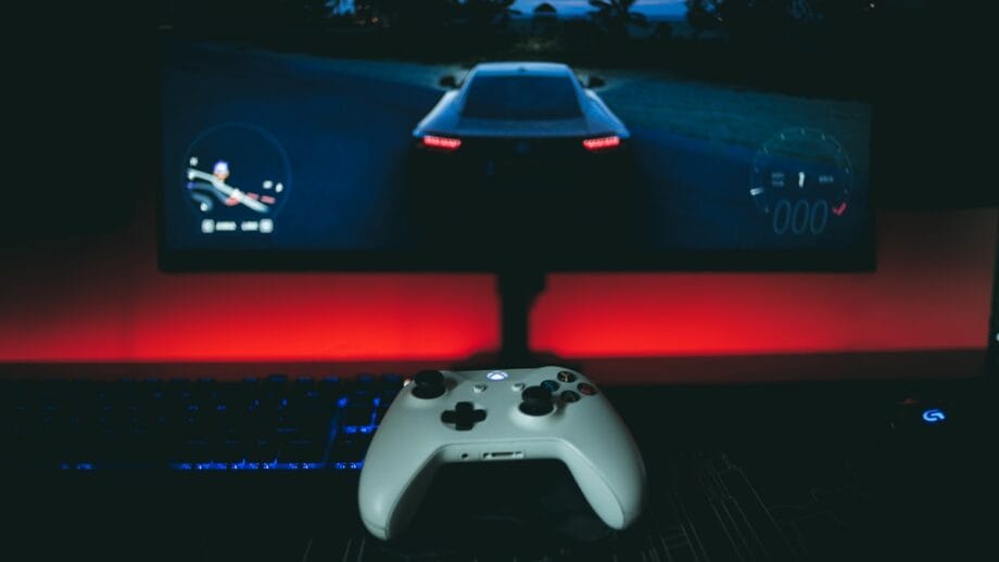 A white game controller sits on a desk in front of a monitor displaying a racing video game with a dim red backlight.