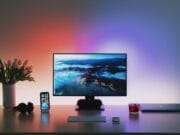 A desk setup featuring a widescreen monitor displaying a scenic landscape, with a wireless keyboard and mouse in front. To the left are white tulips in a vase, an iPhone, and headphones. To the right are a cup of tea and a closed laptop.