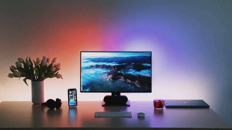 A desk setup featuring a widescreen monitor displaying a scenic landscape, with a wireless keyboard and mouse in front. To the left are white tulips in a vase, an iPhone, and headphones. To the right are a cup of tea and a closed laptop.