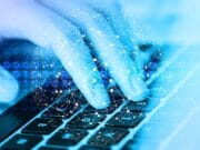 A digital composite image of a human hand touching a laptop keyboard, overlaid with a glowing network of lines and dots, symbolizing connectivity and digital technology.