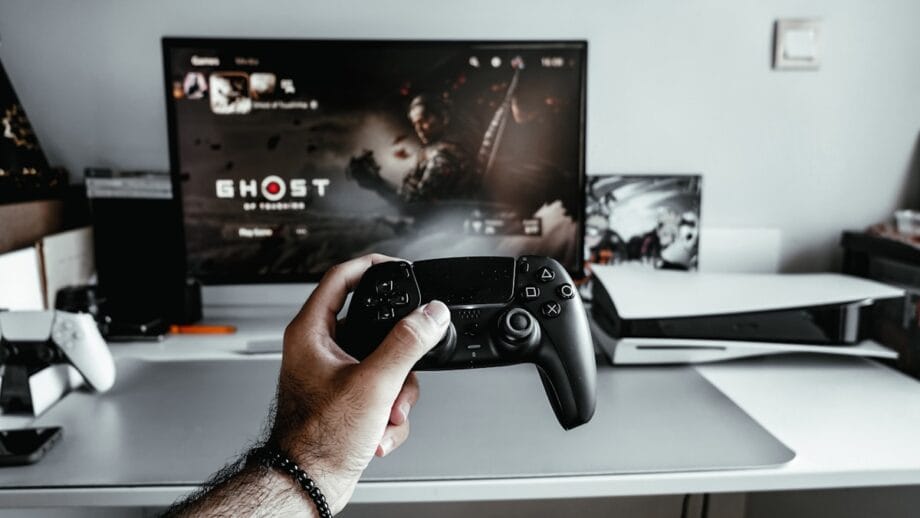 A person holds a black gaming controller in front of a TV screen displaying the game "Ghost of Tsushima" beside a white gaming console on a desk.