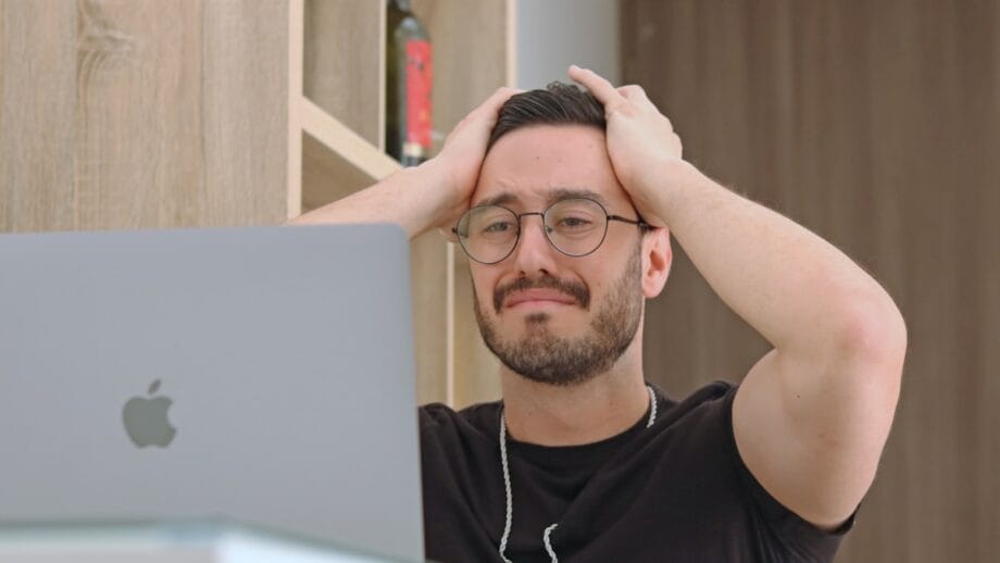A man with glasses looks frustrated while holding his head with both hands in front of a laptop.