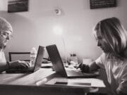 Two people are working on laptops at a table. The person on the left wears a beanie and glasses, while the person on the right has shoulder-length hair. The room is lit by a small desk lamp, and there are various office supplies on the table.
