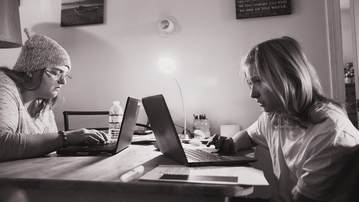 Two people are working on laptops at a table. The person on the left wears a beanie and glasses, while the person on the right has shoulder-length hair. The room is lit by a small desk lamp, and there are various office supplies on the table.