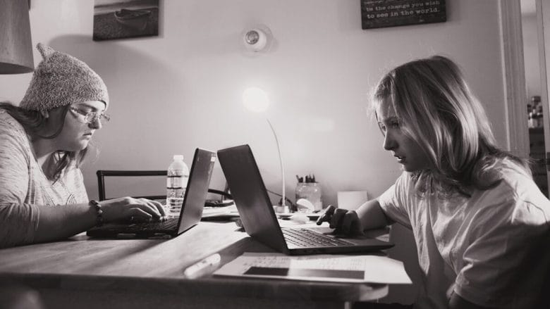 Two people are working on laptops at a table. The person on the left wears a beanie and glasses, while the person on the right has shoulder-length hair. The room is lit by a small desk lamp, and there are various office supplies on the table.