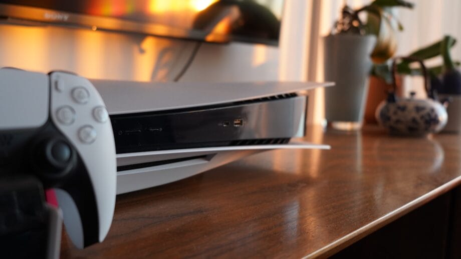 A close-up of a PlayStation 5 console and controller on a wooden surface, with a TV and plants in the background.