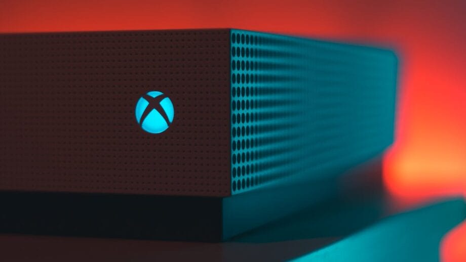 Close-up of a gaming console with a perforated design and an illuminated "X" symbol on the front, set against a background with a red and orange glow.