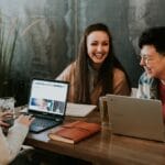 Three people are sitting at a wooden table working on laptops. Two laptops and a notebook are on the table. They are smiling and appear to be engaged in a discussion. A potted plant is in the background against a dark wall.
