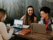 Three people are sitting at a wooden table working on laptops. Two laptops and a notebook are on the table. They are smiling and appear to be engaged in a discussion. A potted plant is in the background against a dark wall.