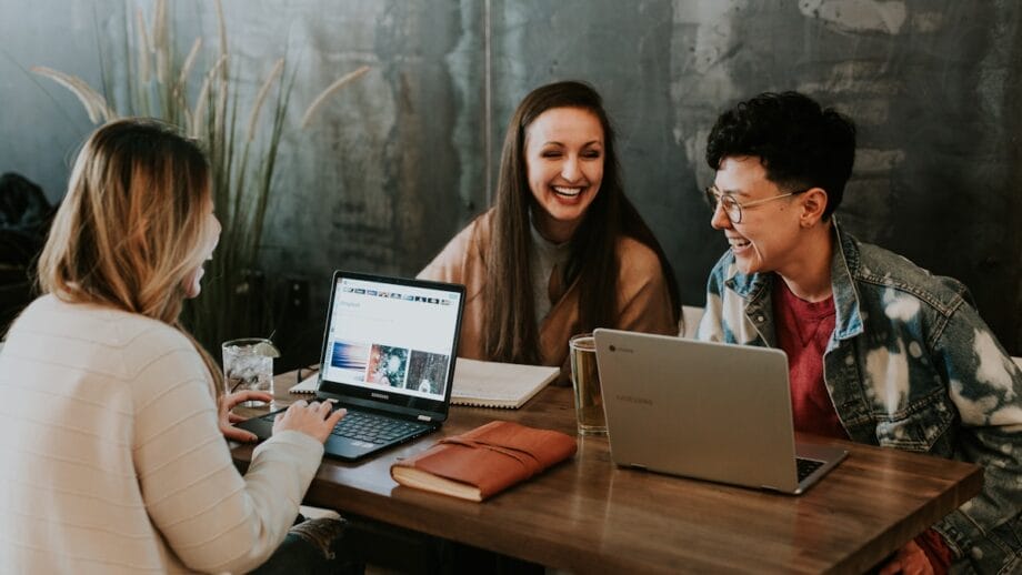 Three people are sitting at a wooden table working on laptops. Two laptops and a notebook are on the table. They are smiling and appear to be engaged in a discussion. A potted plant is in the background against a dark wall.