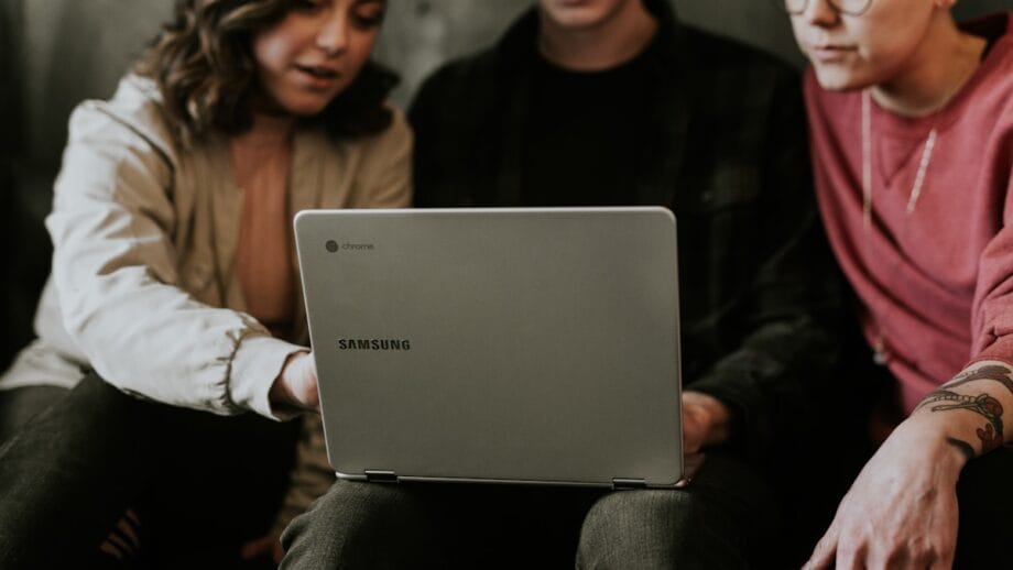 Three people are seated closely together, looking at a silver Samsung Chromebook. One person on the left is pointing at the screen. They appear to be in a casual setting, wearing comfortable clothing.