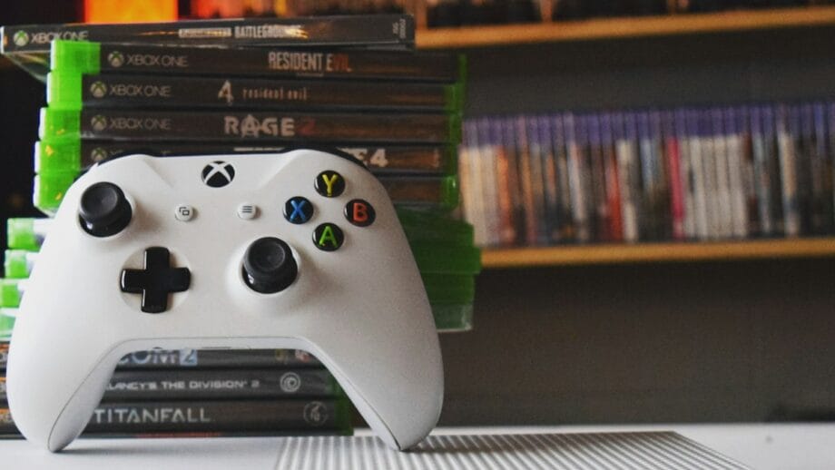 A white Xbox controller is placed in front of a stack of Xbox game cases. Shelves filled with more games or DVDs are visible in the background.