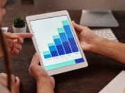 Two people are analyzing a tablet displaying a bar graph with data from January to May. A computer monitor and potted plant are in the background.