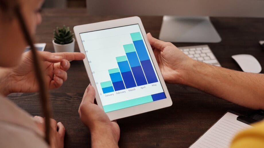 Two people are analyzing a tablet displaying a bar graph with data from January to May. A computer monitor and potted plant are in the background.