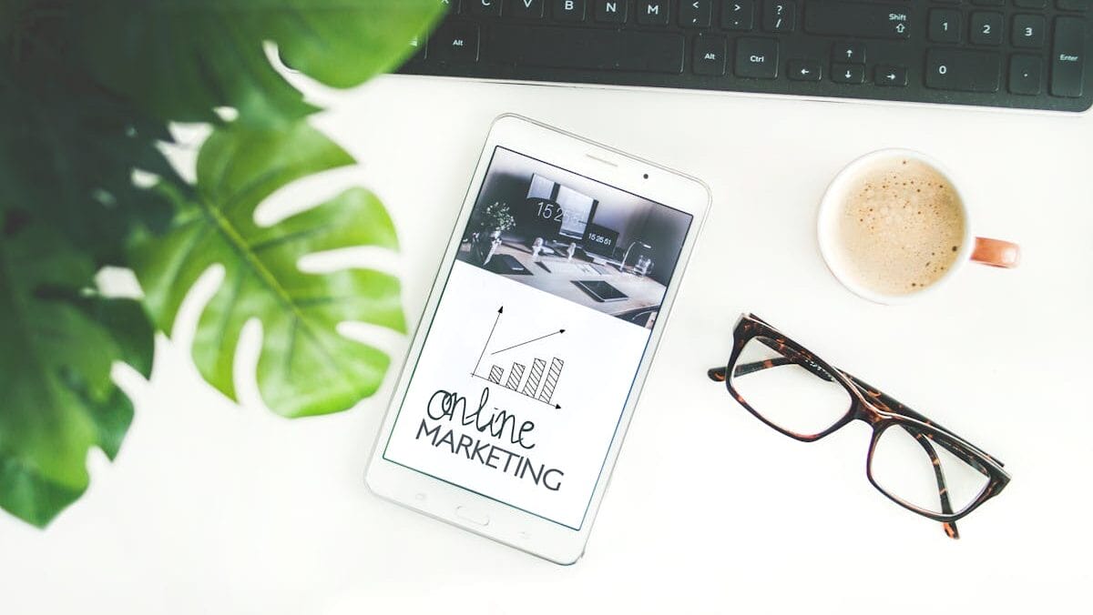 A tablet displaying "online marketing" lies on a white desk next to a plant, glasses, a coffee cup, and a black keyboard.