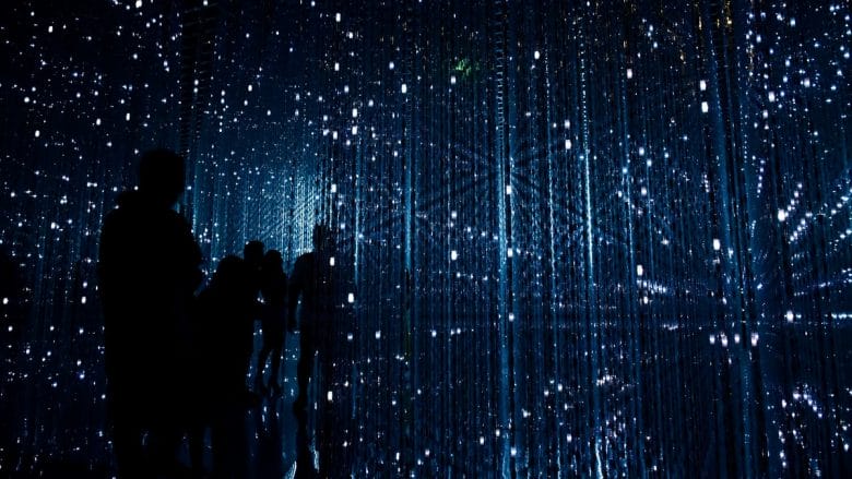 Silhouetted people walk through a dark room filled with vertical, glowing blue light strands.