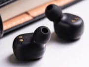 Two black wireless earbuds placed on a surface near a leather wallet or case.