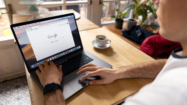 A person using a laptop with the Google search page open on the screen. There is a cup of coffee, plants, and other items on the wooden table in a well-lit room.