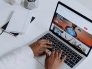 Person typing on a laptop displaying a website, with notebooks and potted plants on a white desk.