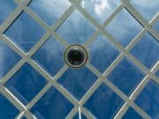A security camera is mounted on a blue glass ceiling with a white lattice structure. The sky and clouds are visible through the glass.