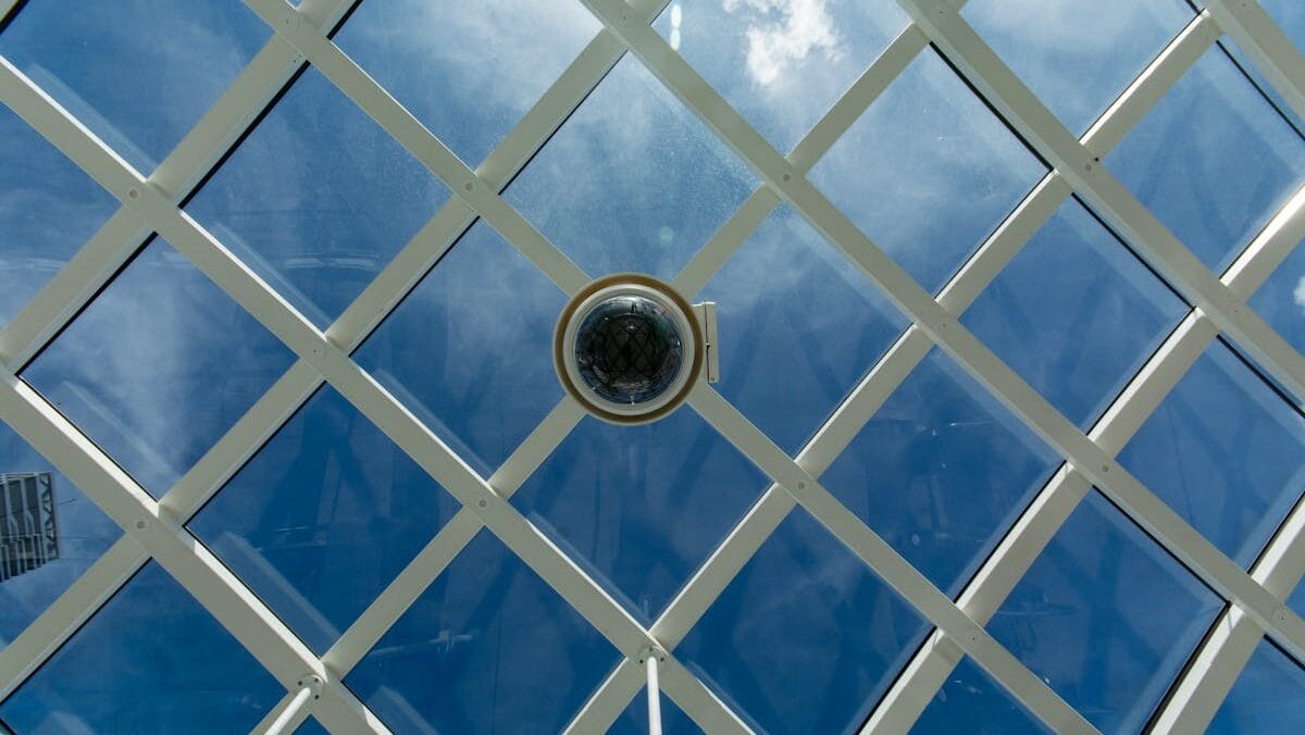 A security camera is mounted on a blue glass ceiling with a white lattice structure. The sky and clouds are visible through the glass.