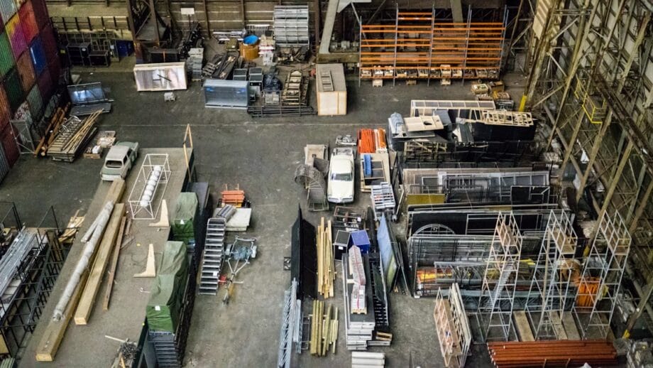 Aerial view of a large warehouse interior with various materials and equipment, including stacked metal beams, wooden pallets, and a white truck, arranged in organized sections.