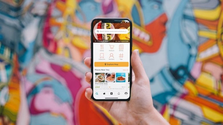 A hand holding a smartphone displaying a food delivery app, with a colorful mural in the background.
