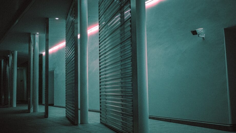 A dimly lit hallway with teal lighting, tall pillars, metal slatted partitions, and a security camera mounted on the wall.