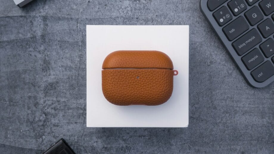 Brown leather case on a white square, surrounded by a keyboard, a tablet, a USB-C hub, a wallet, and a computer mouse on a gray surface.