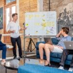 Four individuals in a modern office engage in a meeting. One person presents at a whiteboard while the others listen and sit with laptops in front of them.