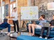 Four individuals in a modern office engage in a meeting. One person presents at a whiteboard while the others listen and sit with laptops in front of them.