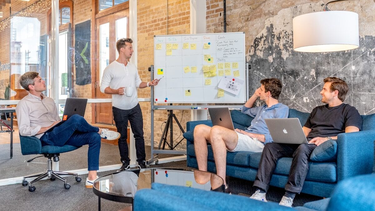 Four individuals in a modern office engage in a meeting. One person presents at a whiteboard while the others listen and sit with laptops in front of them.