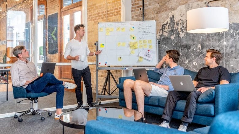 Four individuals in a modern office engage in a meeting. One person presents at a whiteboard while the others listen and sit with laptops in front of them.