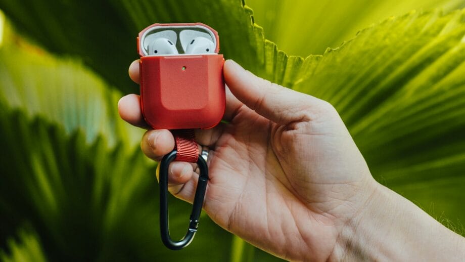 A hand holding a red case with white wireless earbuds in front of large green leaves.