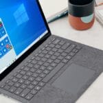 A laptop with a gray keyboard and a Windows 10 start screen displayed. Nearby are a notebook with handwritten notes, a closed notebook, and a colorful mug.