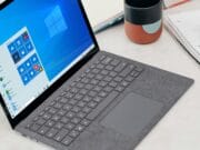 A laptop with a gray keyboard and a Windows 10 start screen displayed. Nearby are a notebook with handwritten notes, a closed notebook, and a colorful mug.