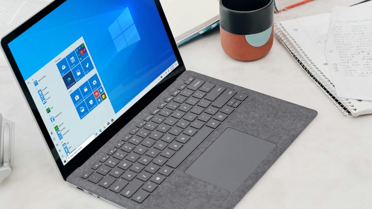 A laptop with a gray keyboard and a Windows 10 start screen displayed. Nearby are a notebook with handwritten notes, a closed notebook, and a colorful mug.