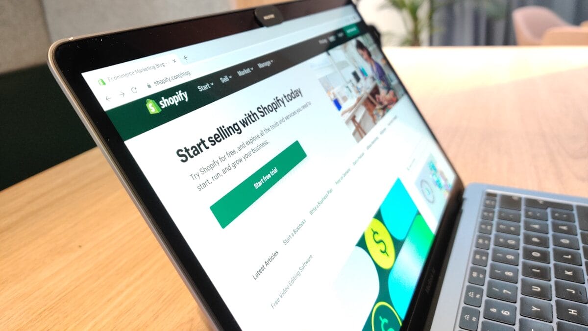 A laptop screen displays the Shopify website, with the text "Start selling with Shopify today" on a page urging users to sign up. The laptop is on a wooden desk beside a plant.