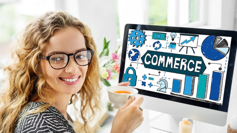 A woman with glasses, smiling and holding a cup, sits at a desk with a computer displaying various commerce-related icons.