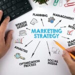 A person works at a desk with a computer keyboard, sketching a diagram labeled "Marketing Strategy" with connected terms like Planning, Management, Social Media, and Optimization Process.