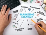 A person works at a desk with a computer keyboard, sketching a diagram labeled "Marketing Strategy" with connected terms like Planning, Management, Social Media, and Optimization Process.