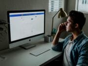 A person sits at a desk, looking at a computer screen displaying a webpage. They are holding a phone to their ear and a lamp is on the desk.