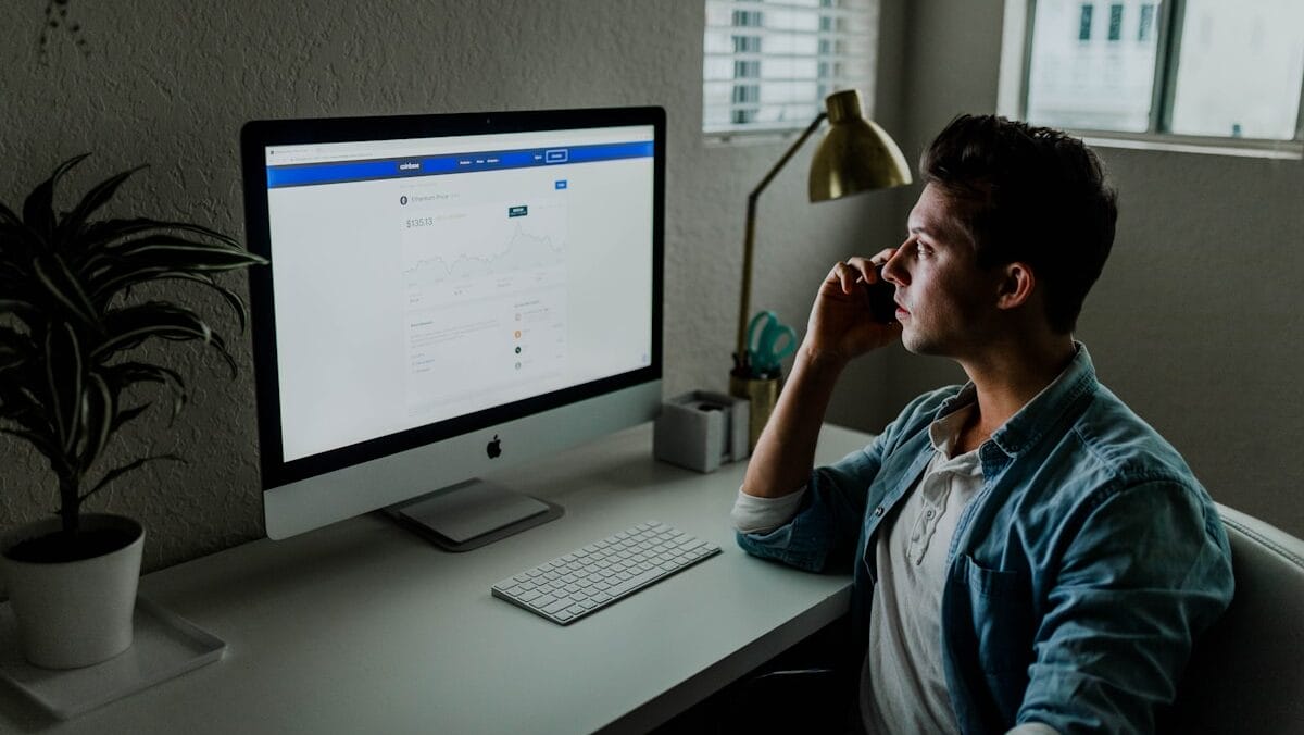 A person sits at a desk, looking at a computer screen displaying a webpage. They are holding a phone to their ear and a lamp is on the desk.