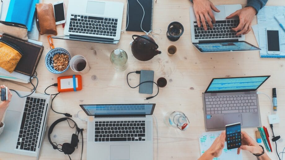 Four people working on laptops around a cluttered table with notebooks, phones, headphones, snacks, and various electronic equipment.