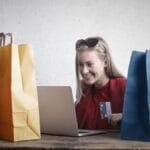 A woman is sitting at a table with multiple shopping bags, using a laptop and holding a credit card.